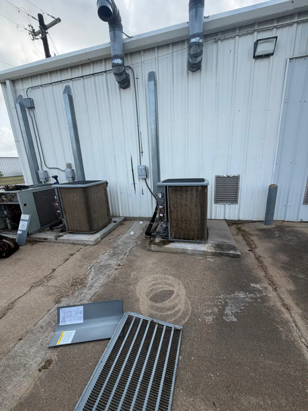 Outdoor condenser units beside a metal building, coils covered in rust and grime, one of them opened up with the wiring exposed and the coil panel lying on the ground